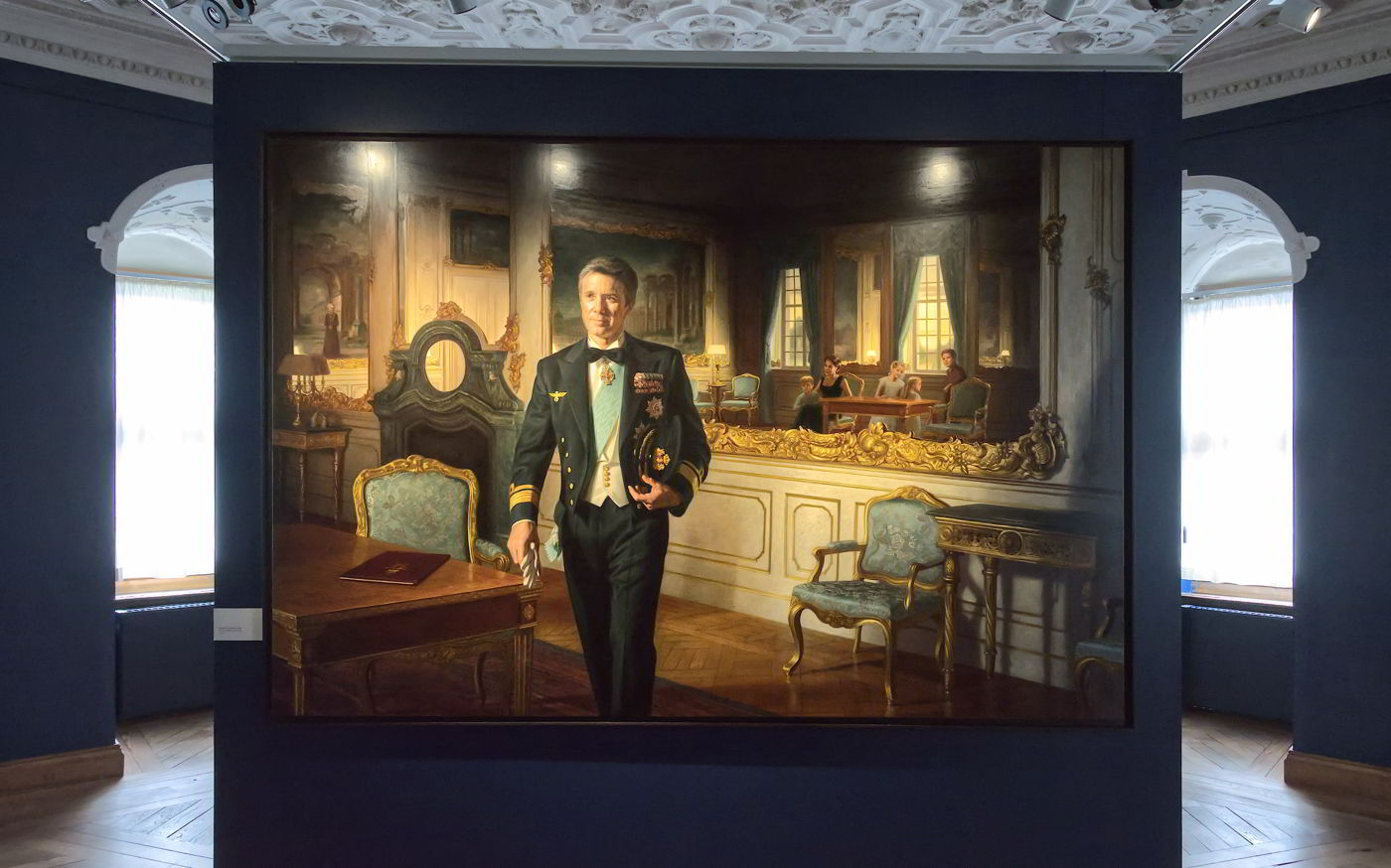 Painting of Crown Prince Frederik in a suit and decorations, hanging on a dark blue wall with spotlight.