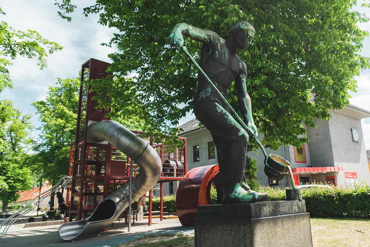 The playground in Frederiksværk with the large slide in the background and a statue of a worker in the foreground.